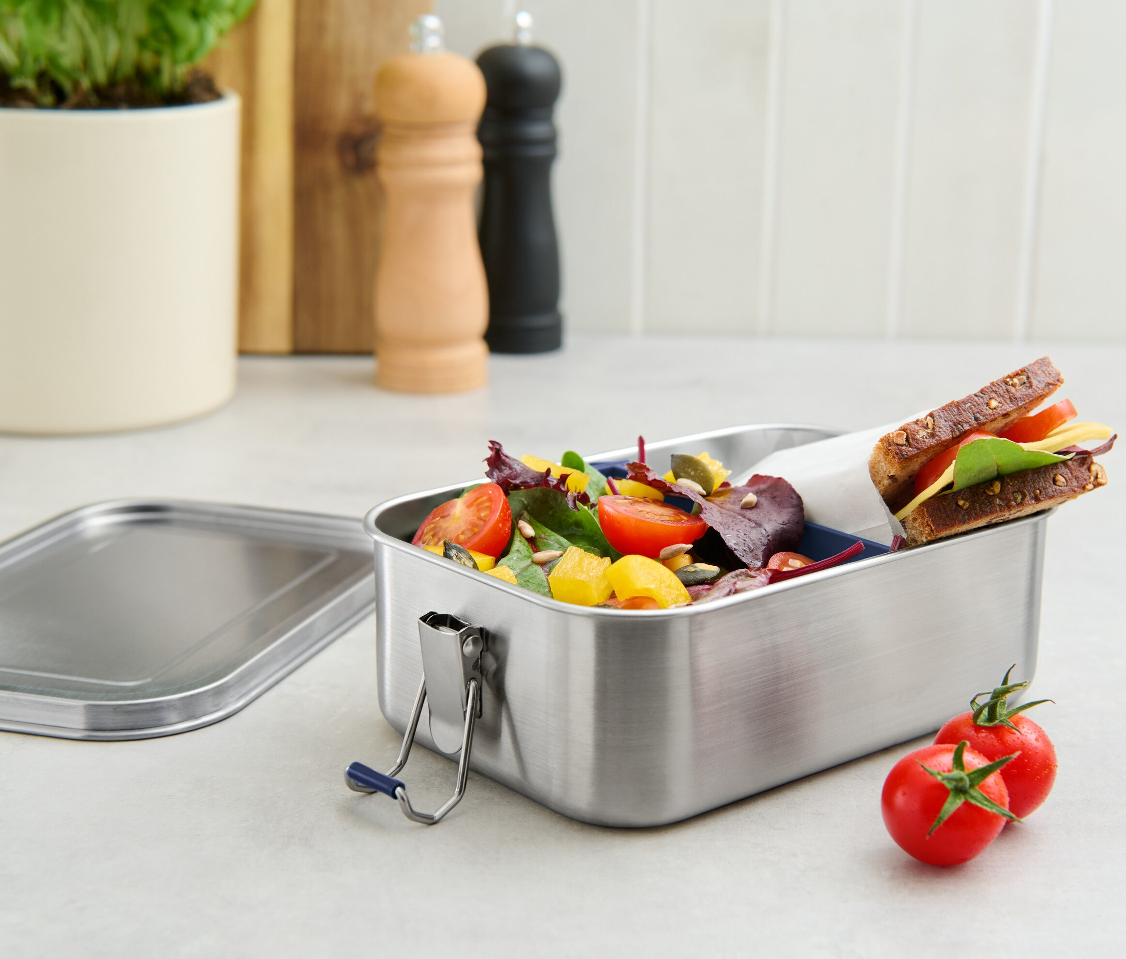 Eine To-go-Lunchbox aus Edelstahl, gefüllt mit Salat und einem Sandwich, daneben liegen zwei Tomaten.