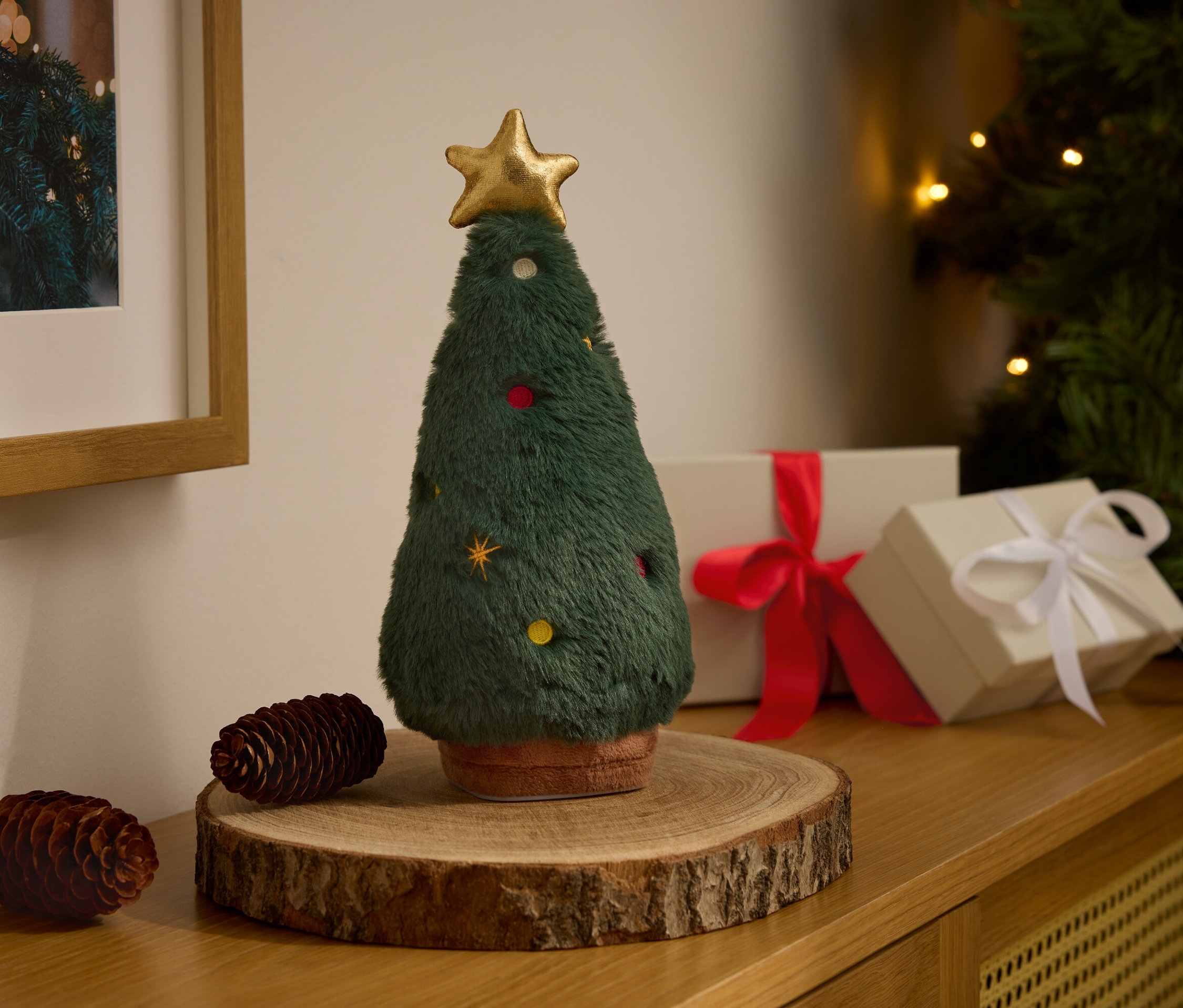 Ein dekorativer Plüsch-Weihnachtsbaum mit goldenem Stern steht auf einem Holzbock neben zwei Tannenzapfen und Geschenkboxen.