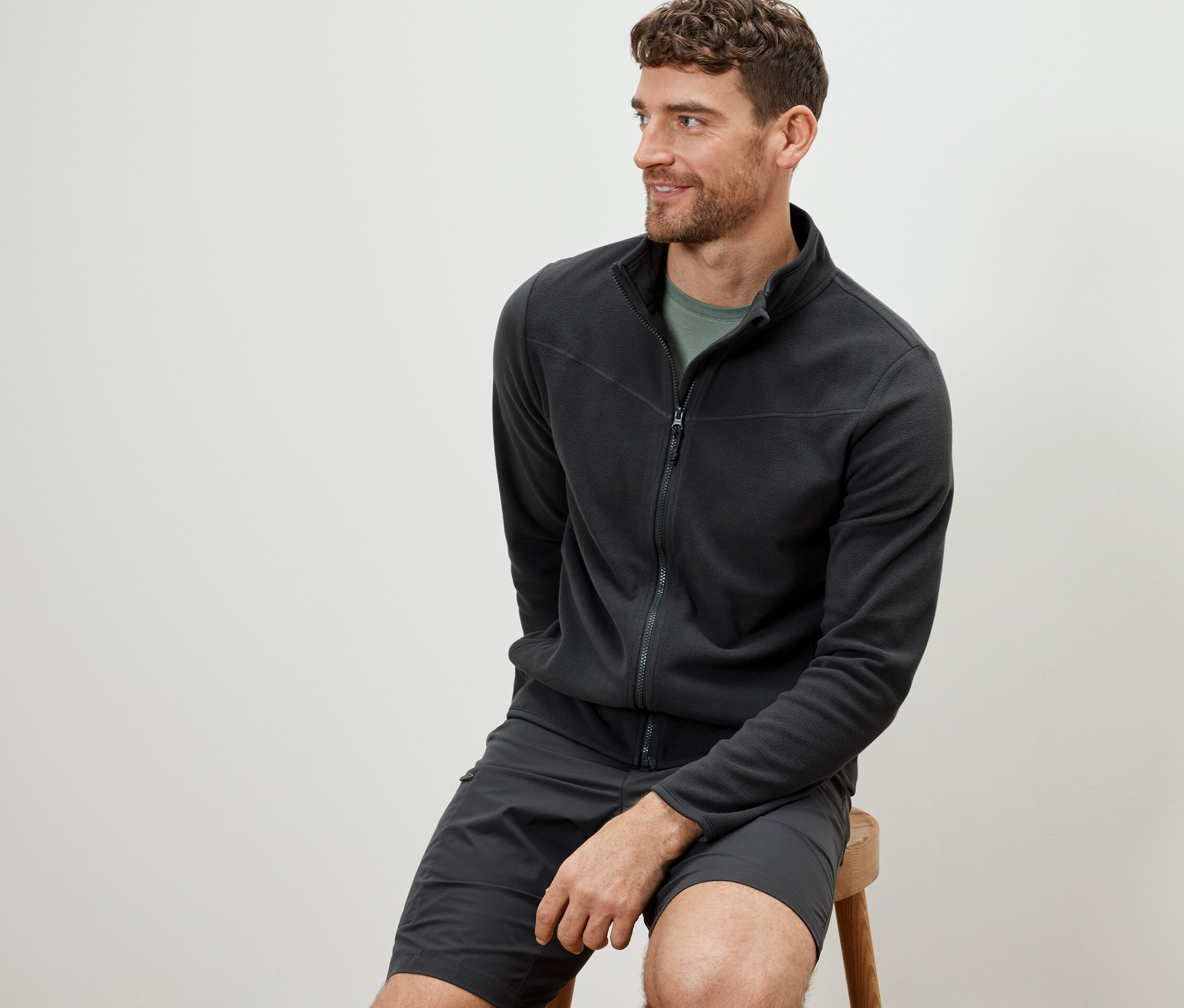 Mann sitzt auf einem Hocker und trägt eine anthrazitfarbene Fleecejacke.