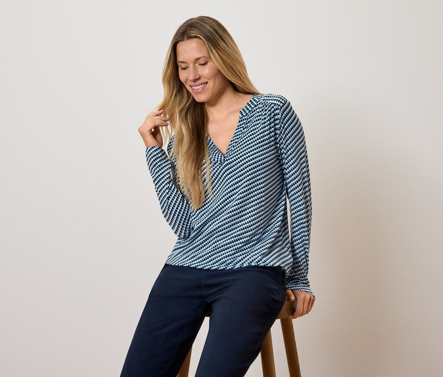 Frau sitzt auf einem Hocker, trägt ein blau-weiß gestreiftes Langarmshirt und dunkelblaue Hosen.