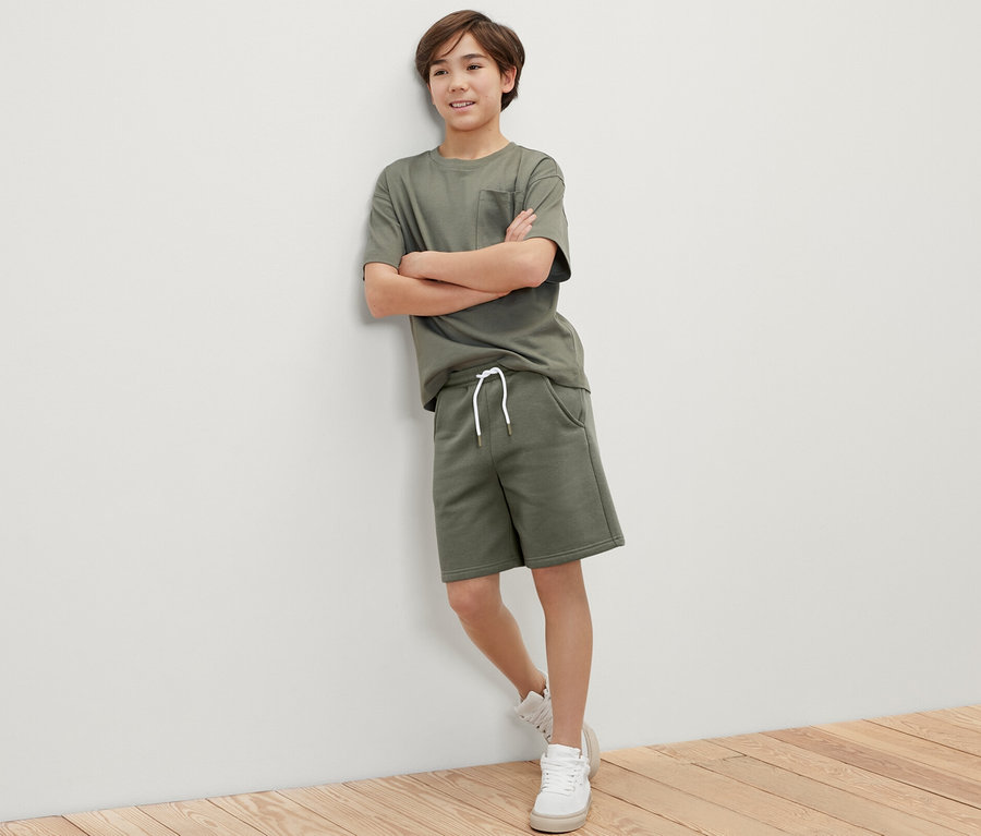 Junge in khaki-farbenem T-Shirt- und Sweatshorts-Set, 2 Kinder-Sweatshorts, steht an einer Wand.