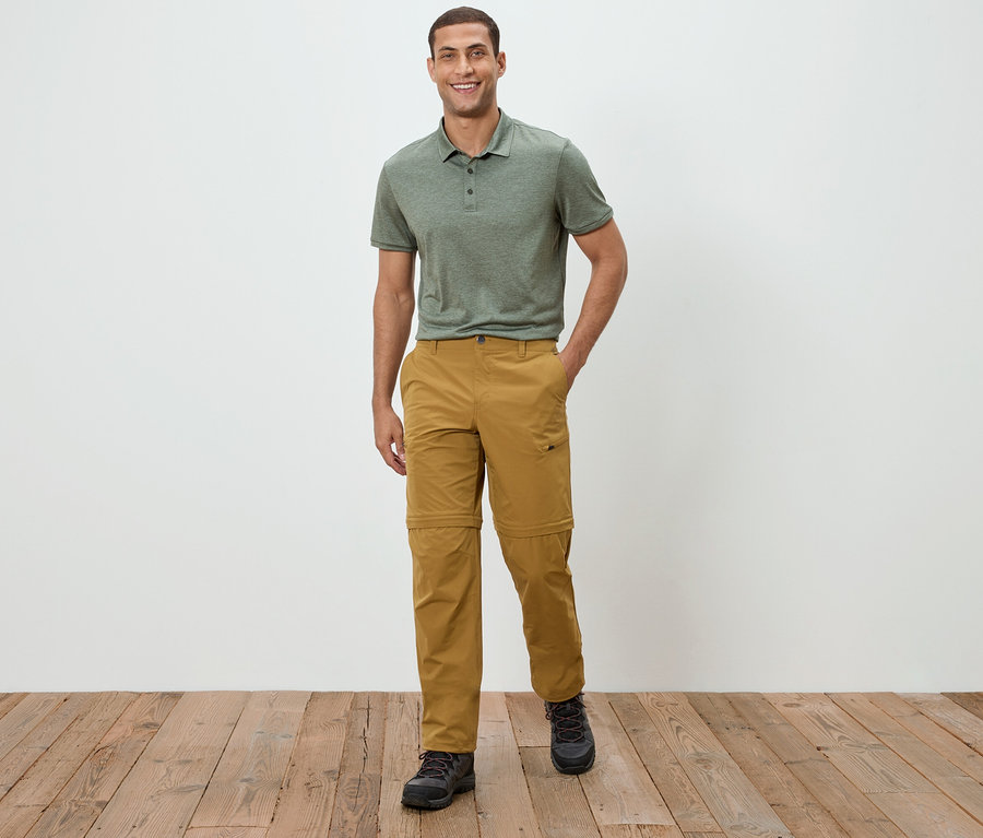 Ein Mann steht vor einer weißen Wand und trägt ein grünes Polohemd und Khakihosen.