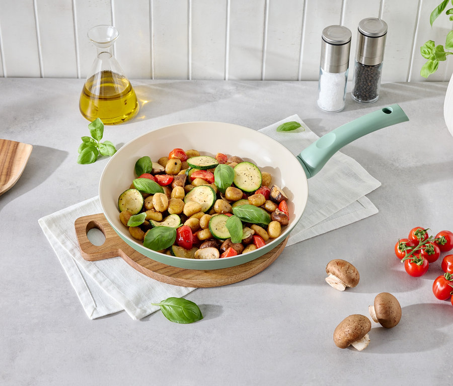 Gemüse-Gnocchi in einer hellgrünen Pfanne auf einem Holzteller. Daneben liegen Champignons, Tomaten, eine Holzkelle und eine Flasche Öl.