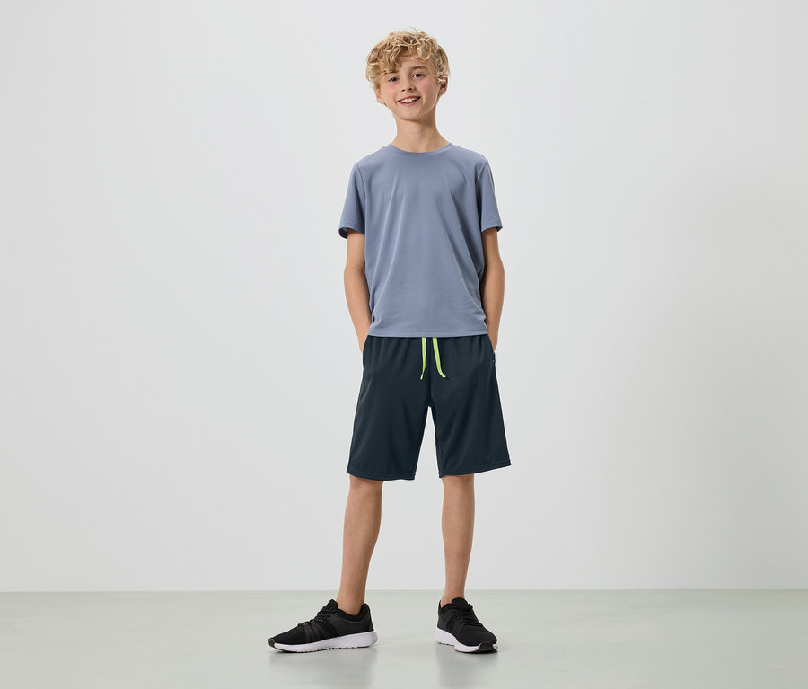 Ein junger Mann mit blonden Locken steht in einem grauen T-Shirt, dunkelblauen Shorts und schwarzen Turnschuhen vor einem weißen Hintergrund.