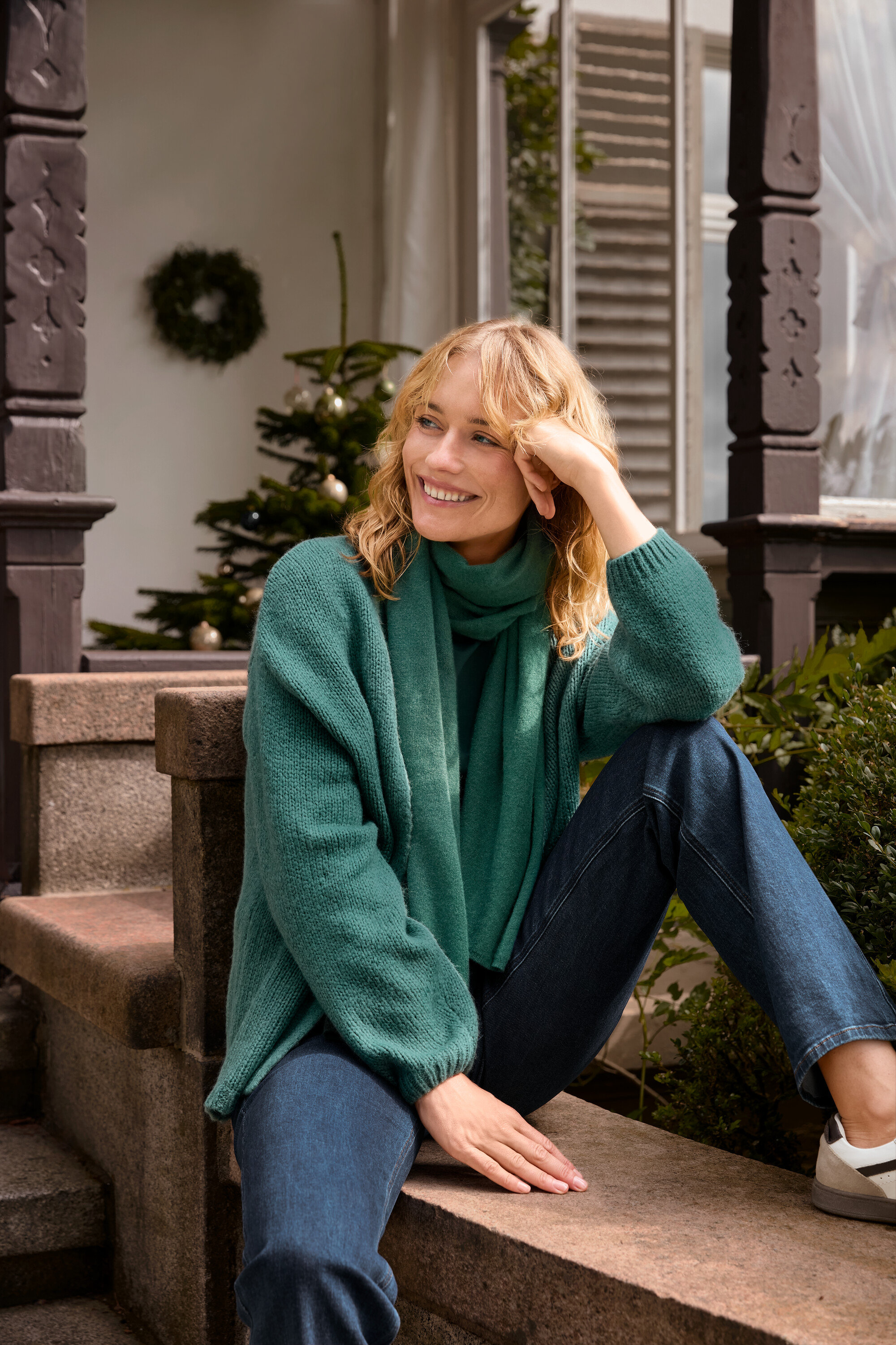 Blonde Frau sitzt auf einer Treppe und trägt einen offenen Grobstrick-Cardigan, einen grünen Webschal und eine Bootcut Jeans – Fit »Lou«.