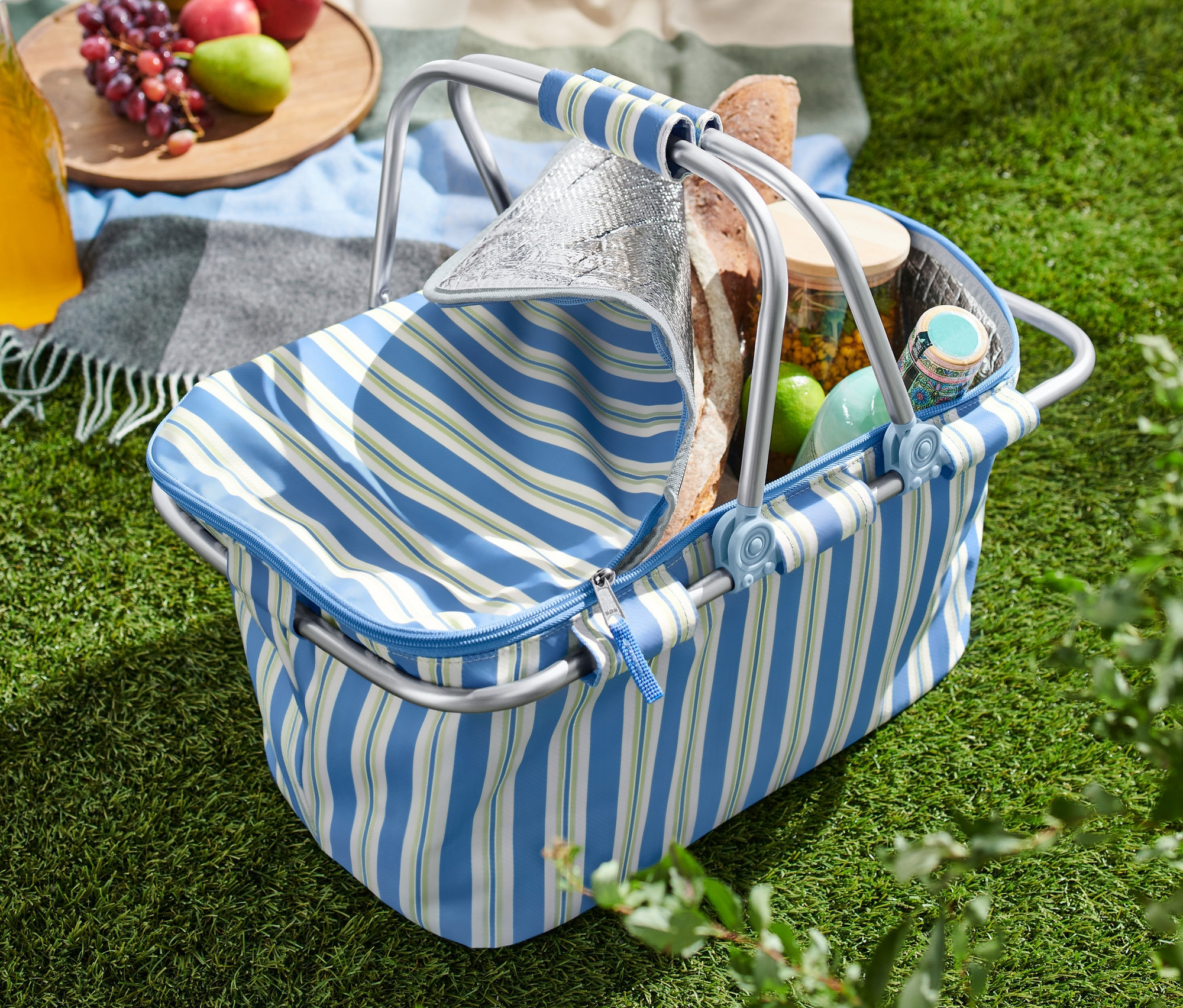 Ein offener Kühlkorb steht auf einer Wiese, daneben eine Decke mit Äpfeln, Trauben und einem Getränk.