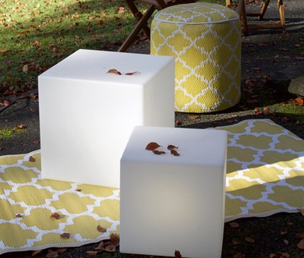 Zwei weiße 8 seasons design LED-Leuchtwürfel, 43 cm, stehen auf einem gelb-weißen Outdoor-Teppich. Dahinter befinden sich ein gelb-weißer Pouf und ein Holztisch mit Stühlen.