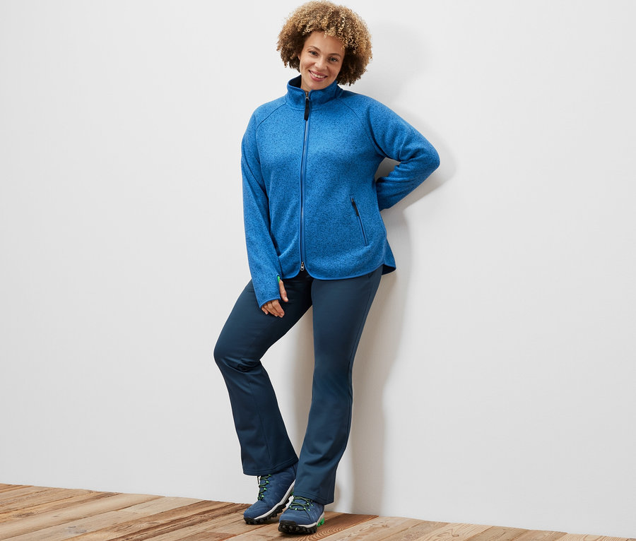 Frau posiert an einer Wand, sie trägt eine blaue Strickfleece-Jacke und Thermo-Jazzpants.