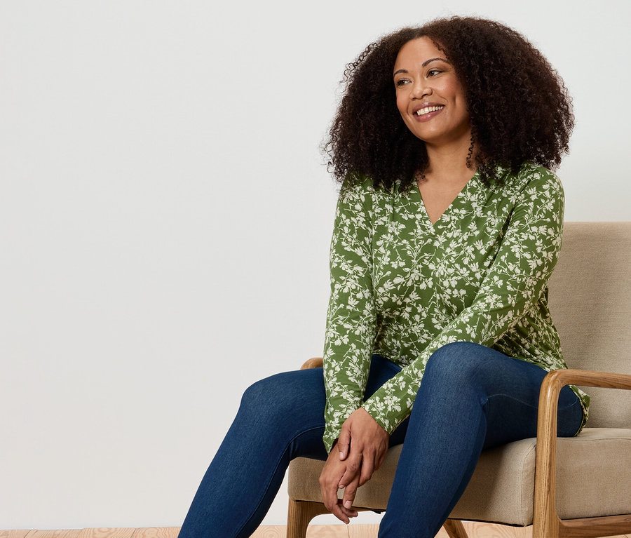 Eine Frau sitzt in einem Sessel. Sie trägt eine grüne Bluse mit Blumenmuster und Jeans.