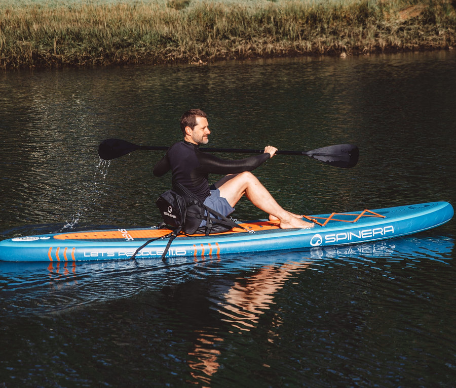 Mann sitzt auf einem blauen Spinera SUP Board Lets Paddle 11' 6" auf ruhigem Wasser und hält ein Paddel.