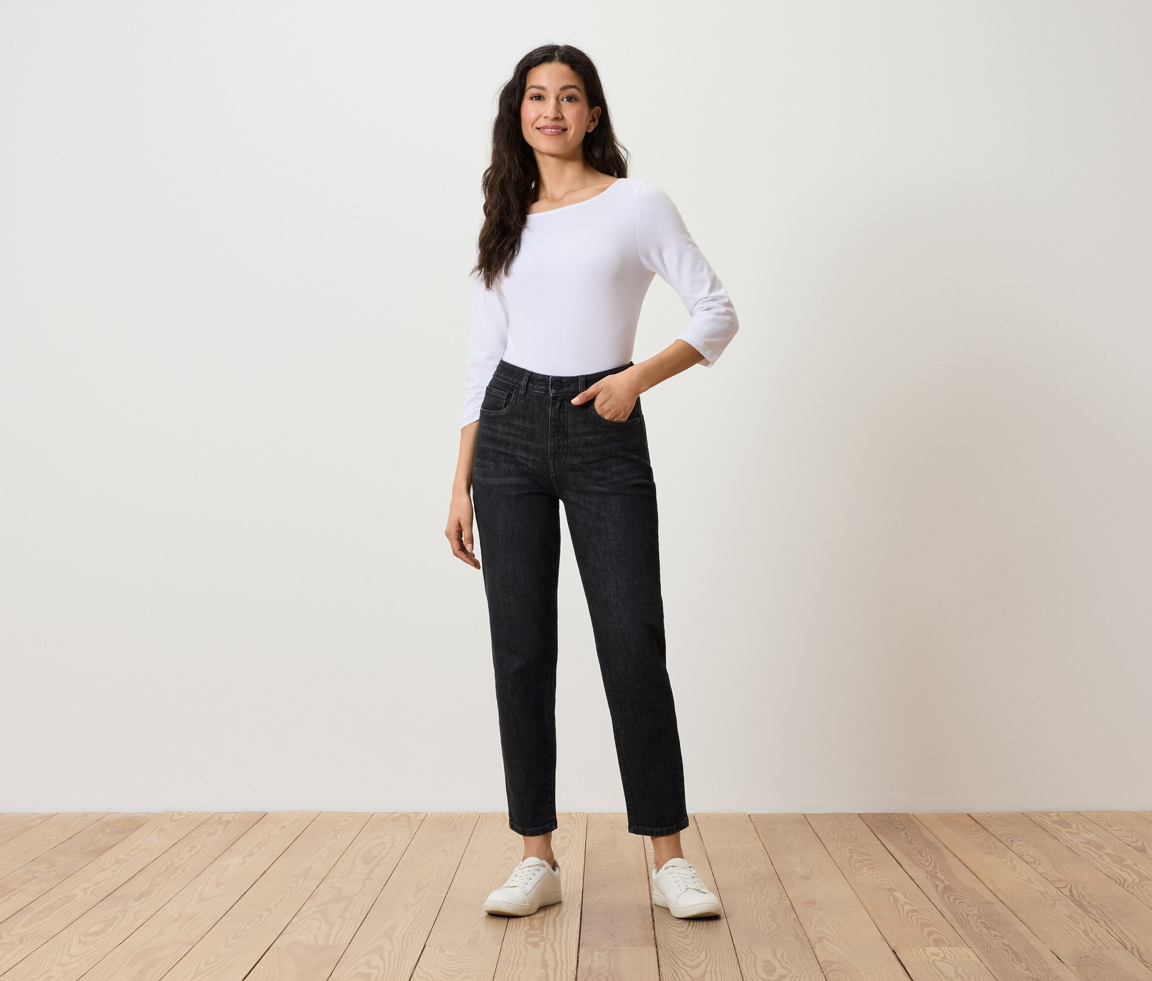 Frau posiert in schwarzen Jeans, weißem Dreiviertelarm-Top und weißen Turnschuhen.