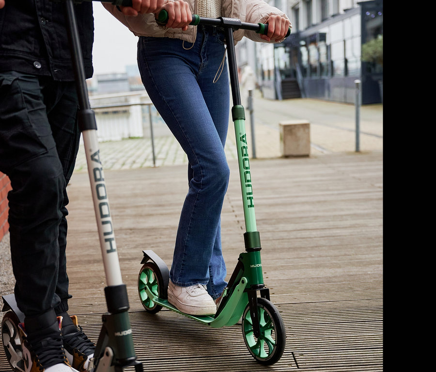 Zwei Personen fahren auf HUDORA BigWheel® 205 advanced Rollern, einer grün, einer grau-weiß, auf einem Holzweg.