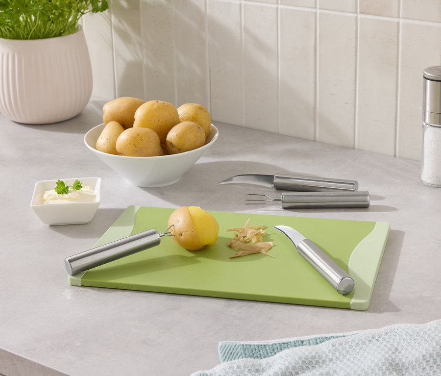 Kartoffeln in einer Schüssel, eine Kartoffel wird auf einem grünen Brett geschält.