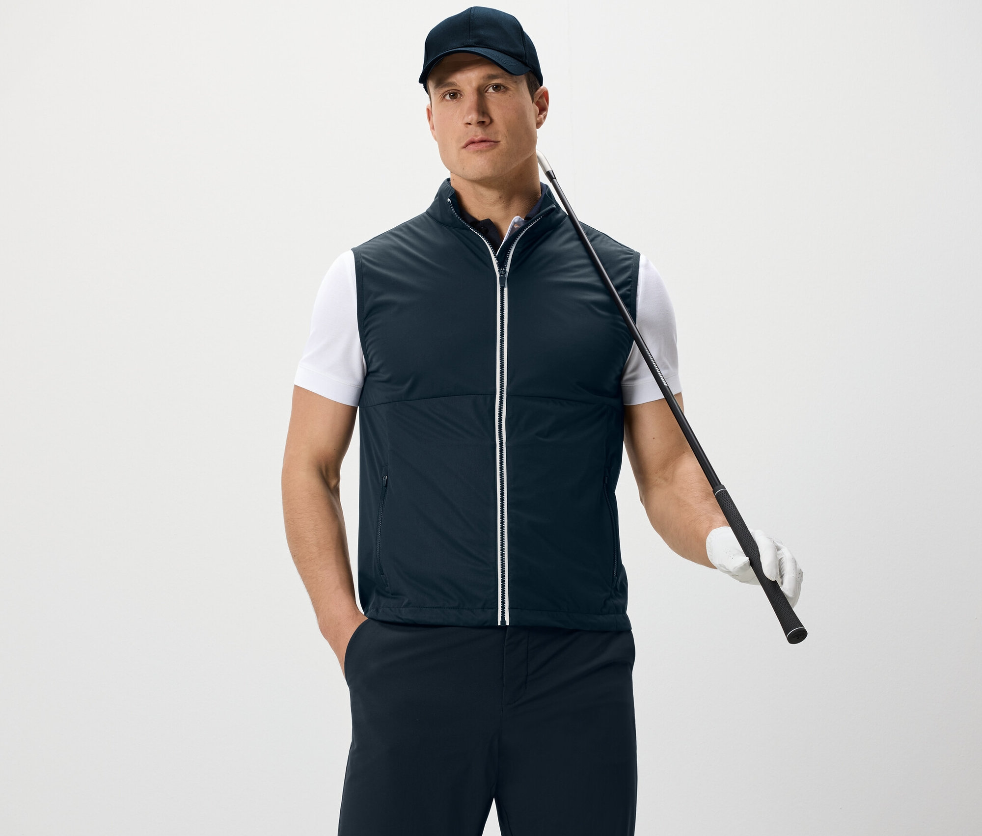 Ein Mann trägt eine Softshellweste und eine Kappe, hält einen Golfschläger und steht vor einem weißen Hintergrund.
