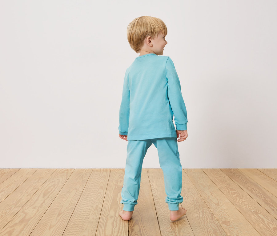 Kleiner Junge von hinten, trägt hellblauen Schlafanzug, steht auf Holzboden vor einer weißen Wand.