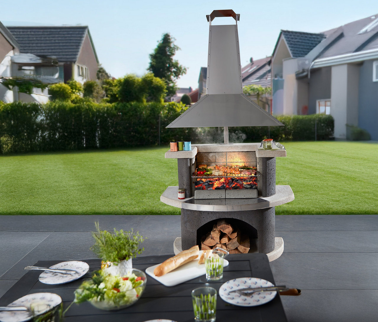 Ein gemauerter Außengrill steht auf einer Terrasse mit einem gedeckten Tisch im Vordergrund.