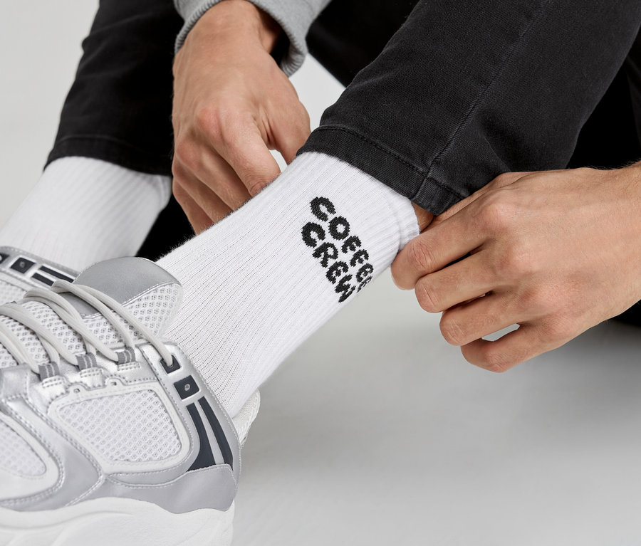 Mann zieht weiße Socken mit der Aufschrift „Coffee Crew“ und silberne Turnschuhe an.