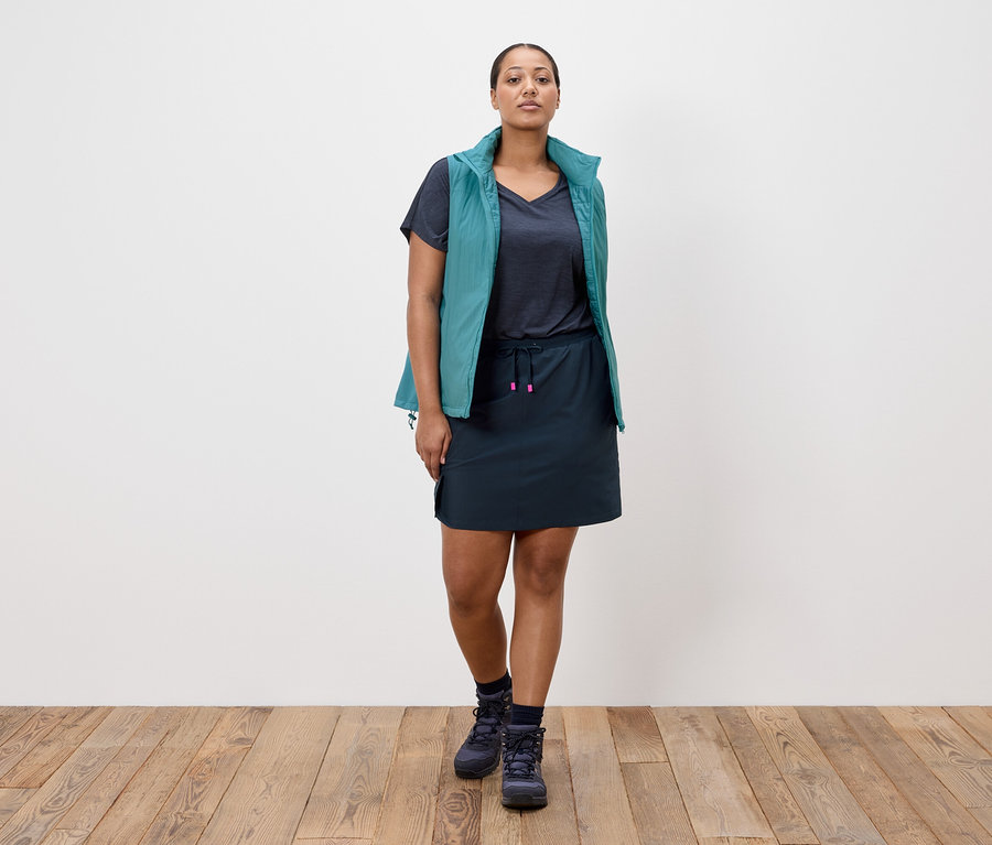 Frau steht vor einer weißen Wand, trägt einen marineblauen Rock, ein marineblaues T-Shirt, eine petrolfarbene Weste und marineblaue Wanderschuhe.