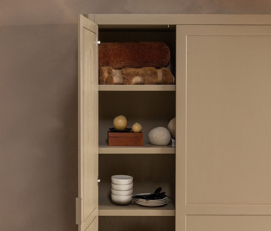 Offener beiger Schrank mit braunen Handtüchern, Dekokugeln, einer Holzschatulle, Schalen und Tellern.