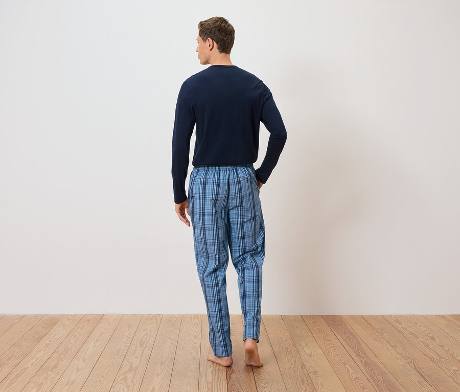 Mann von hinten in blau karierter Schlafanzughose und langärmligem Oberteil vor weisser Wand auf Holzboden.