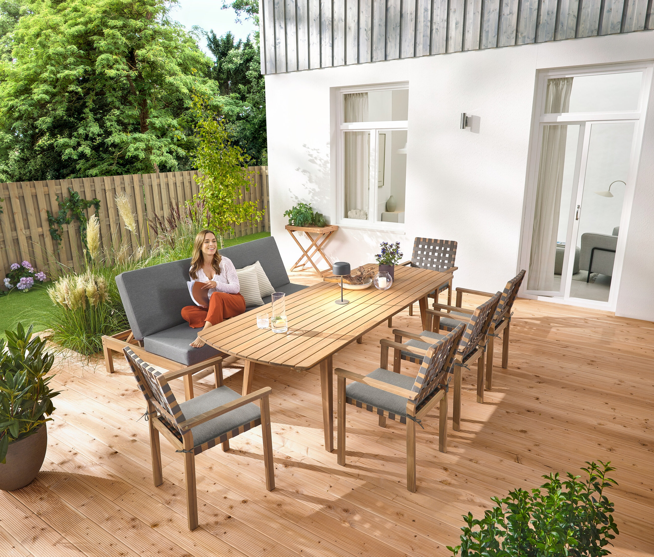 Eine Frau sitzt auf einer 2-in-1-Gartenbank ‒ Doppel-Sonnenliege. Auf dem Ausziehbaren Gartentisch mit abgerundeten Fronten, ca. 180‒240 cm steht eine graue Solar-Akku-Tischleuchte. Es gibt auch einen Garten-Tabletttisch und Stapelbare Gartenstühle mit Gurtgeflecht.