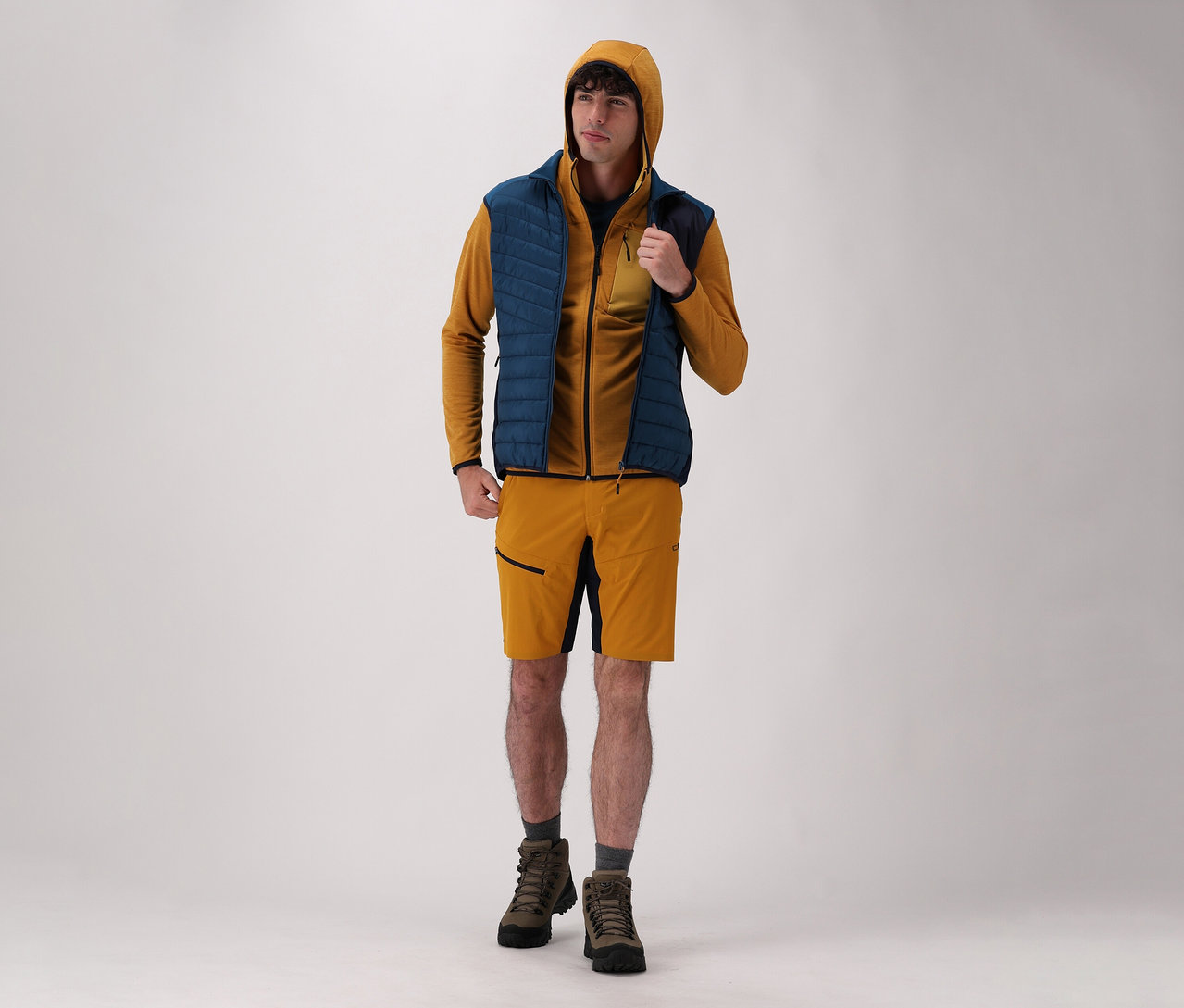 Mann trägt CMP Herren-Kapuzen-Jacke, CMP Herren-Hybridweste, Shorts und Stiefel vor einem hellen Hintergrund.
