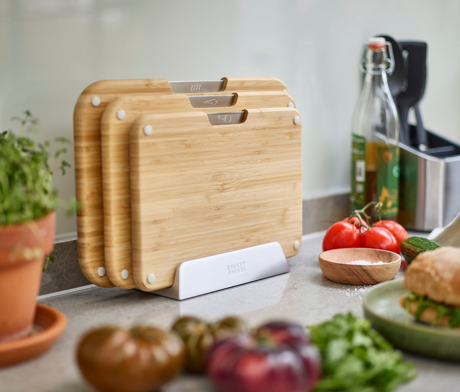 Drei Holzschneidebretter mit silbernen Griffen stehen in einem weißen Halter auf einer Küchentheke, umgeben von Gemüse und Kräutern.