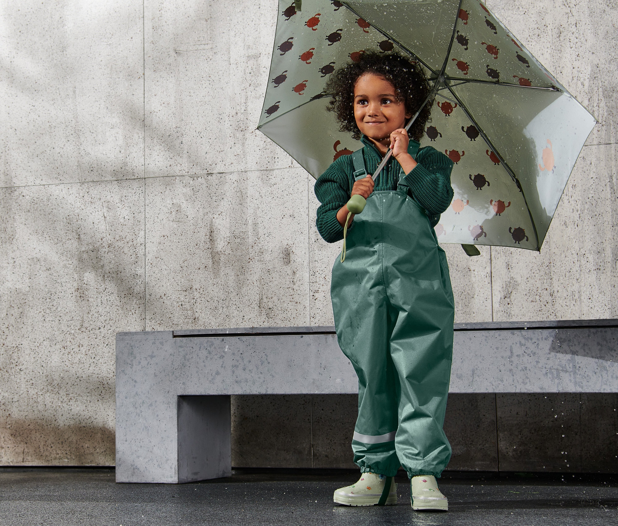 Mädchen hält einen Schirm mit Schildkrötenmuster. Sie trägt eine Kinder-Thermo-Regenhose, dunkelgrün.