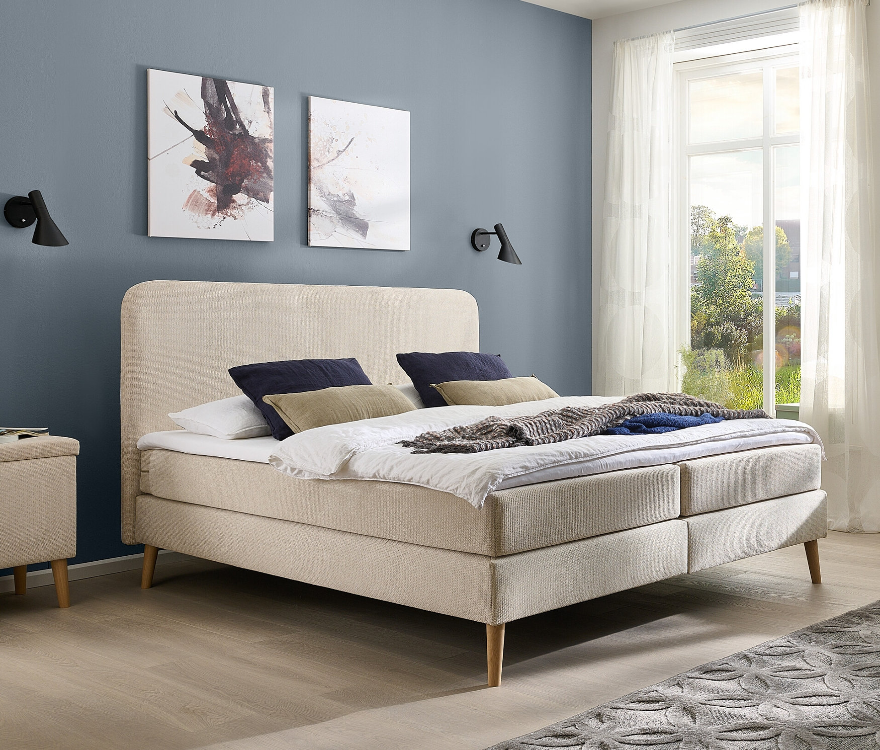 Schlafzimmer mit beigem DOMO collection Boxspringbett mit festem Kopfteil, ca. 180 x 200 cm, inkl. Topper, zwei Nachttischen und grauem Teppich.