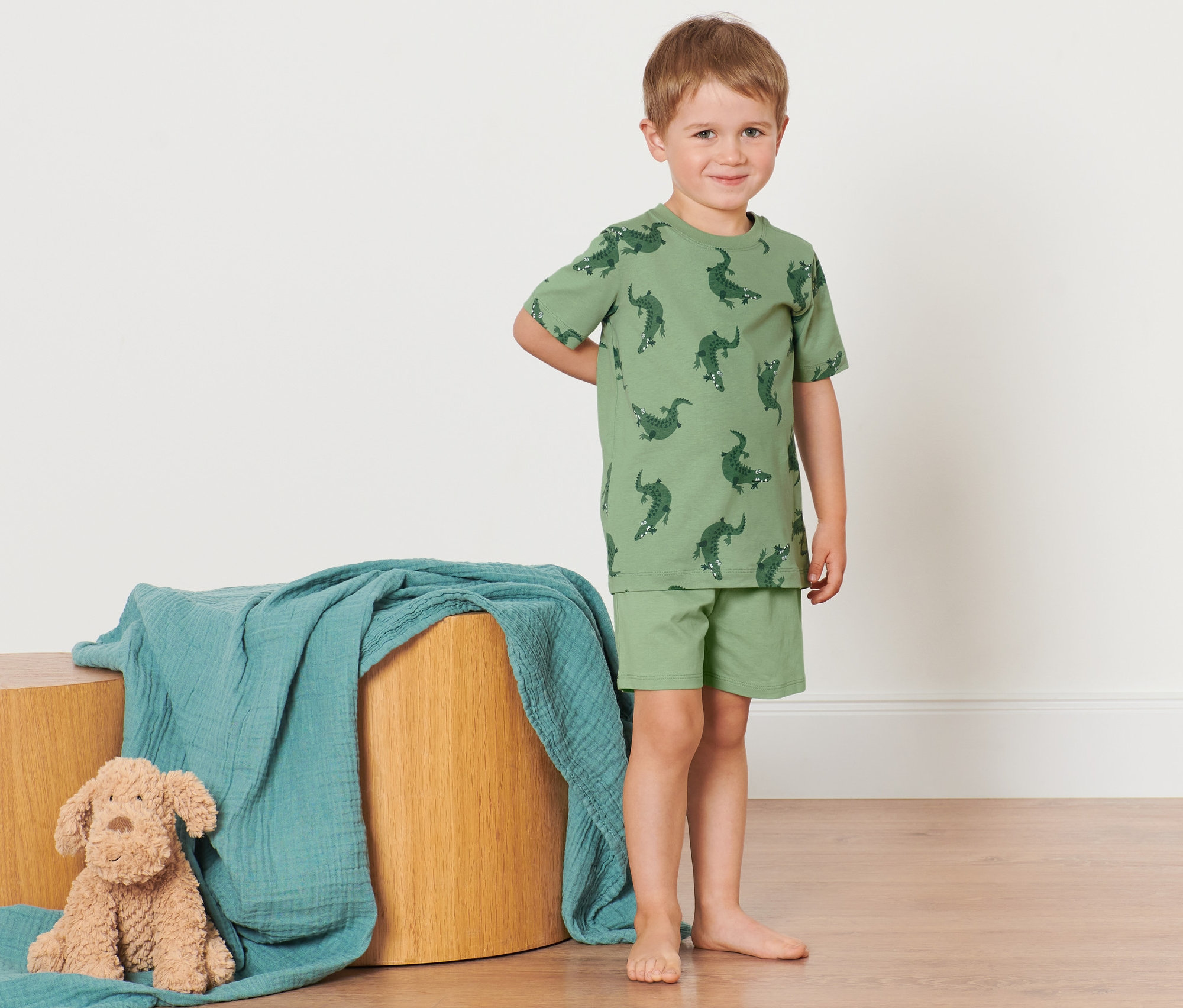 Ein Junge im Kinder-Shorty-Pyjama mit Krokodilmuster steht neben einem Holztisch mit einer blauen Decke und einem Stoffhund.