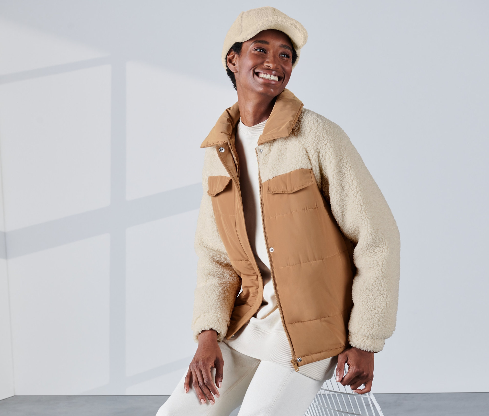 Eine lächelnde Frau sitzt und trägt eine cognacfarbene und weiße Steppjacke mit Teddy-Akzenten und eine Baskenmütze.