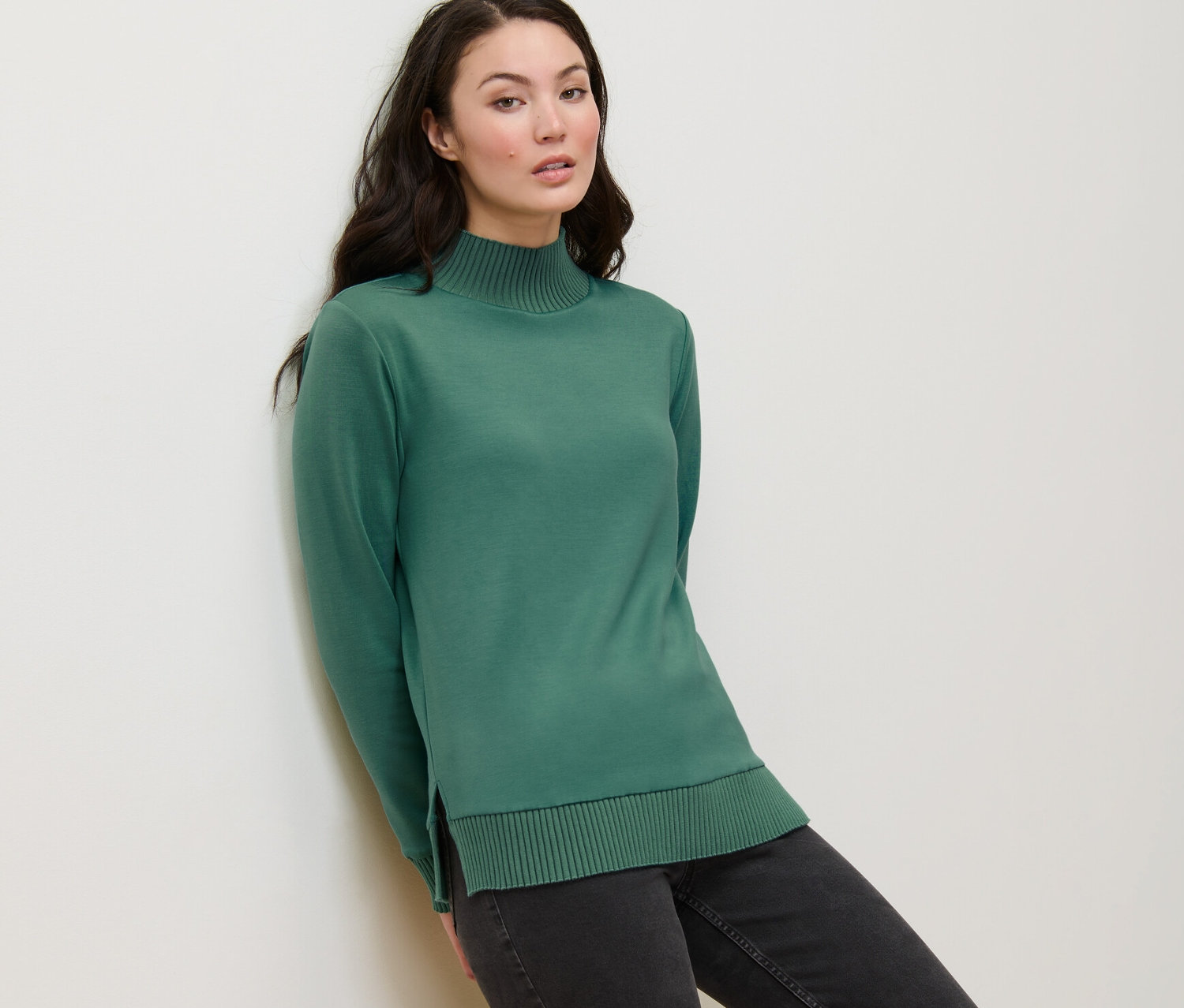 Eine Frau lehnt an einer Wand und trägt ein grünes Sweatshirt mit Stehkragen.