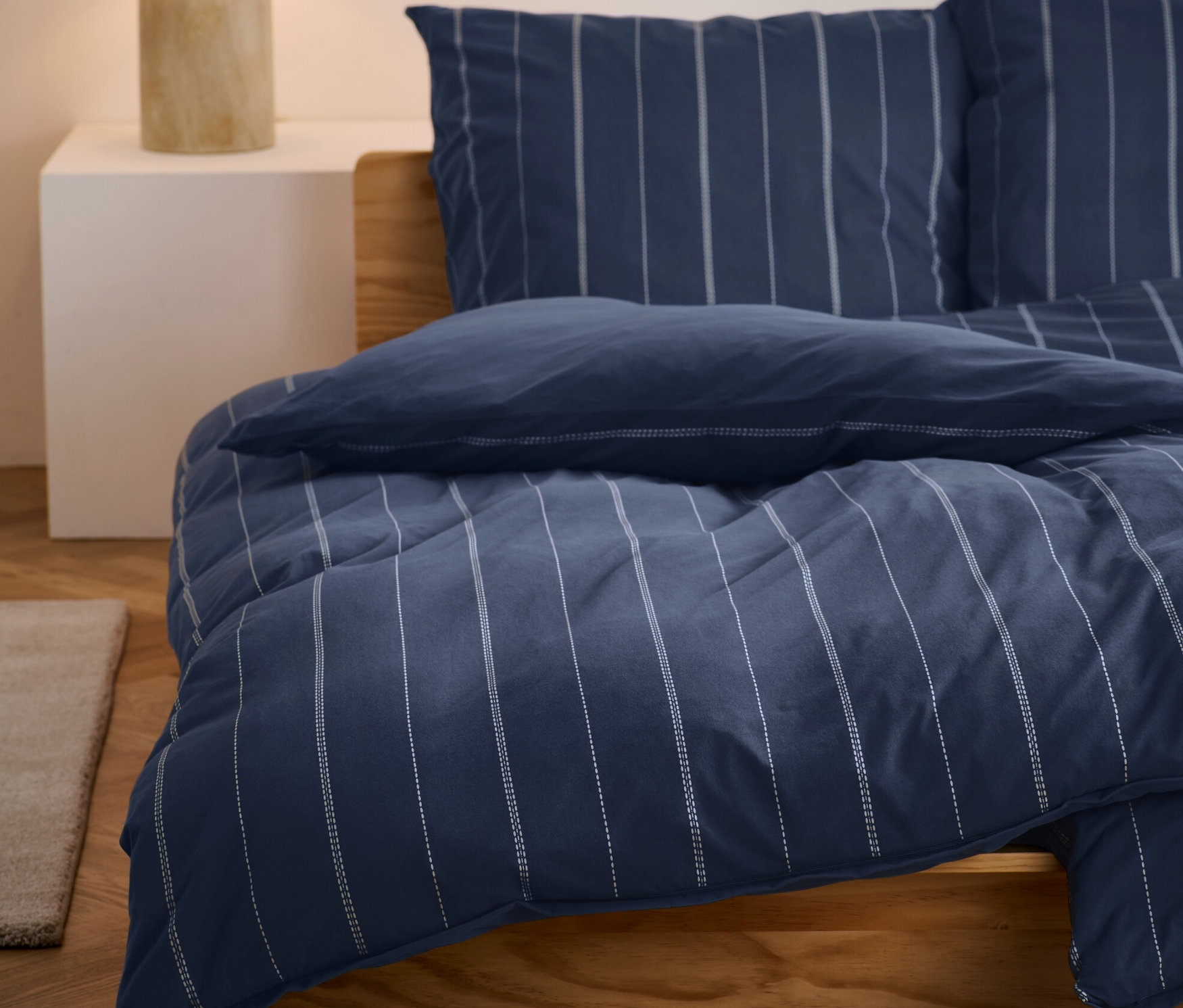 Blau gestreifte Winterjersey-Bettwäsche liegt auf einem Bett. Auf einem Nachttisch steht eine Lampe.