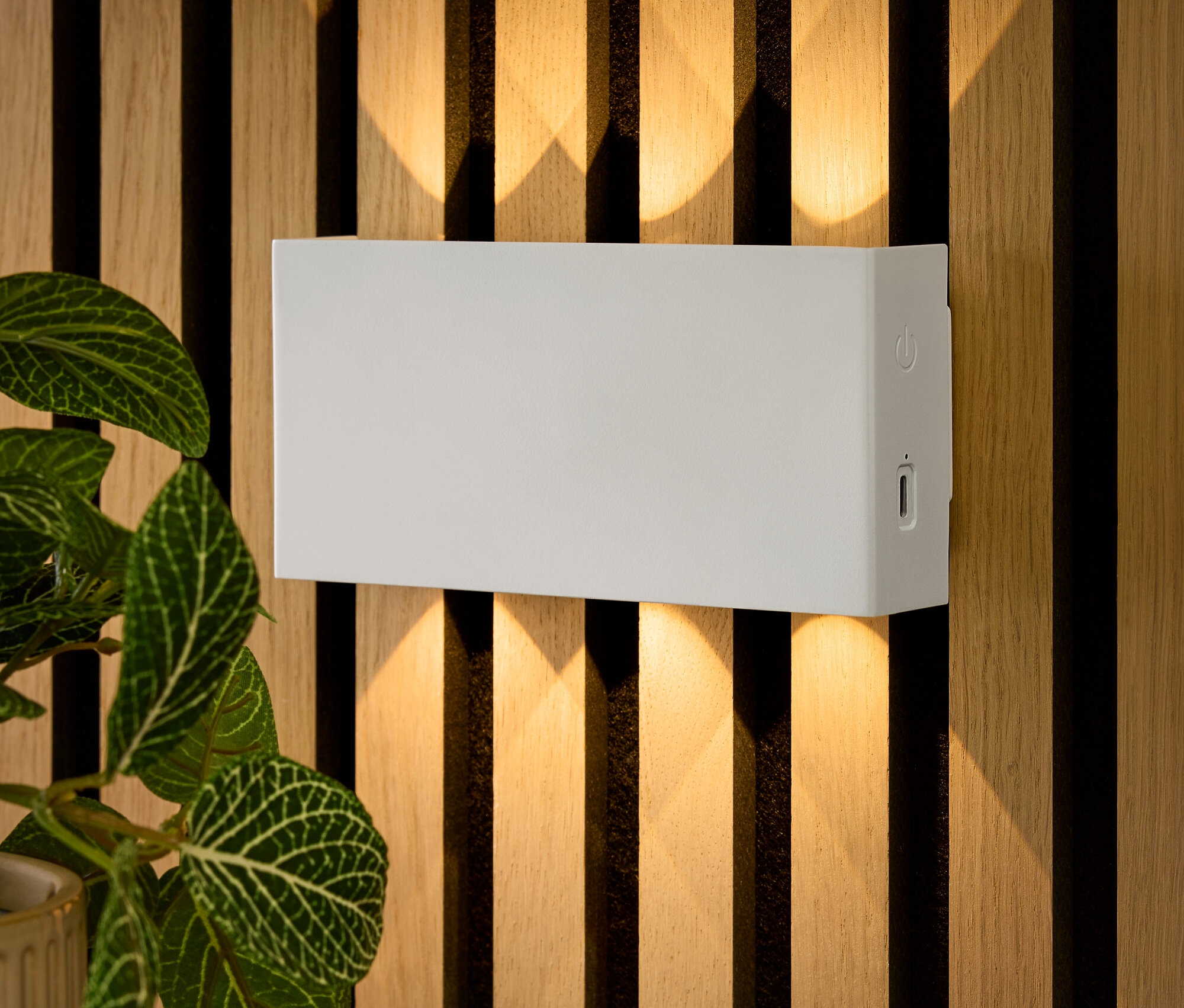 Eine rechteckige, weiße Wandleuchte ist an einer Holzwand befestigt. Darunter liegen Schlüssel auf einem Tisch und eine Blume steht in einem Topf.
