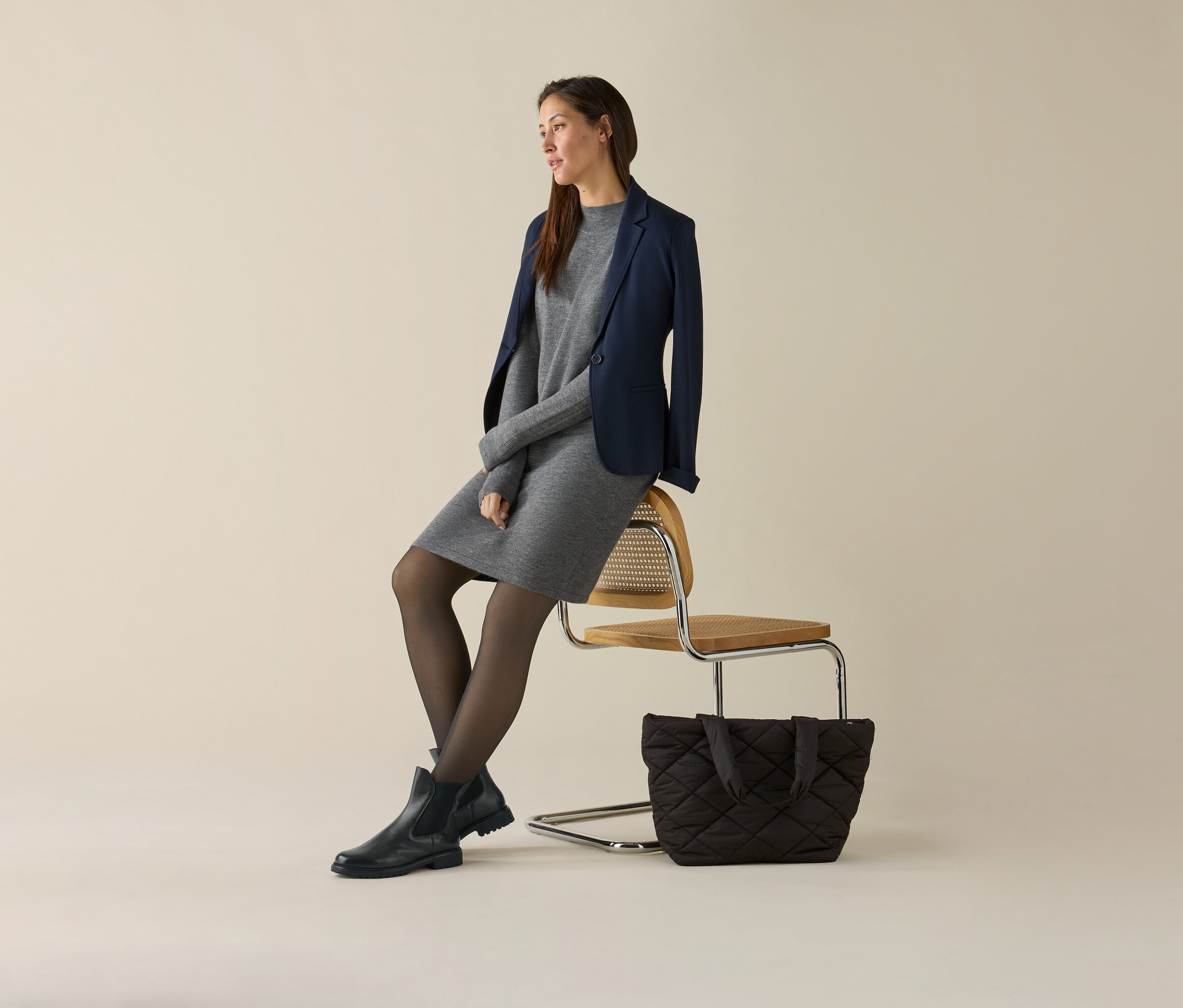 Frau sitzt auf einem Stuhl, trägt ein graues Kleid und einen marineblauen Jerseyblazer. Neben dem Stuhl steht eine Handtasche.