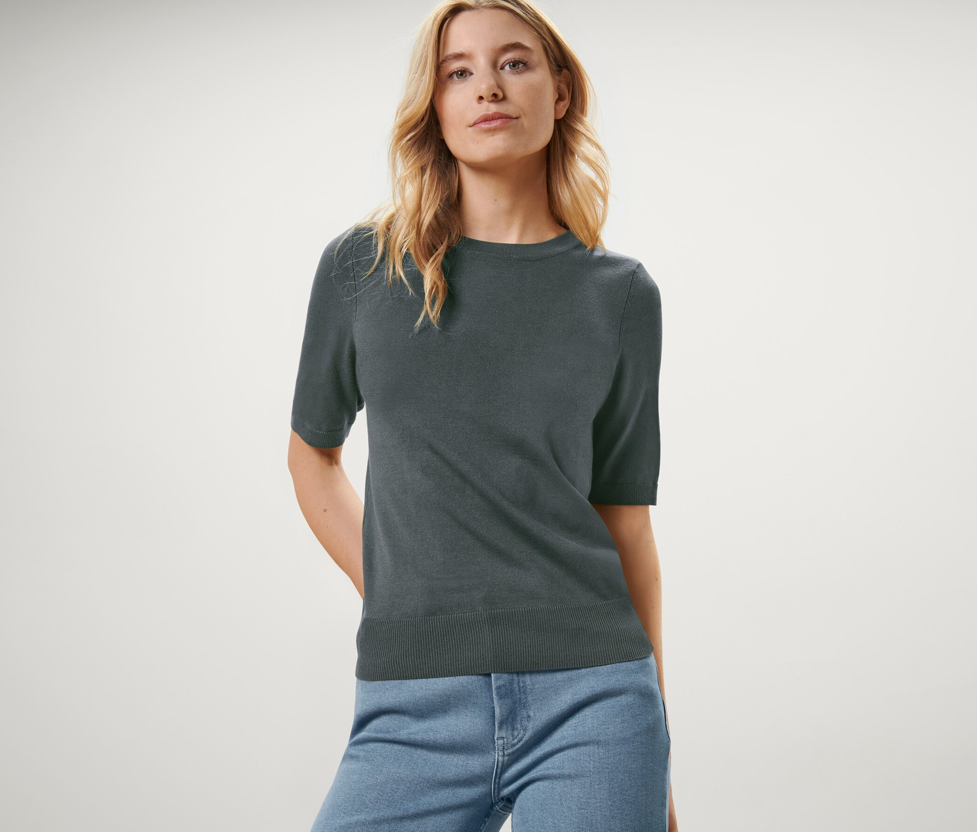 Frau posiert in einem grünen Feinstrickpullover und blauen Jeans.