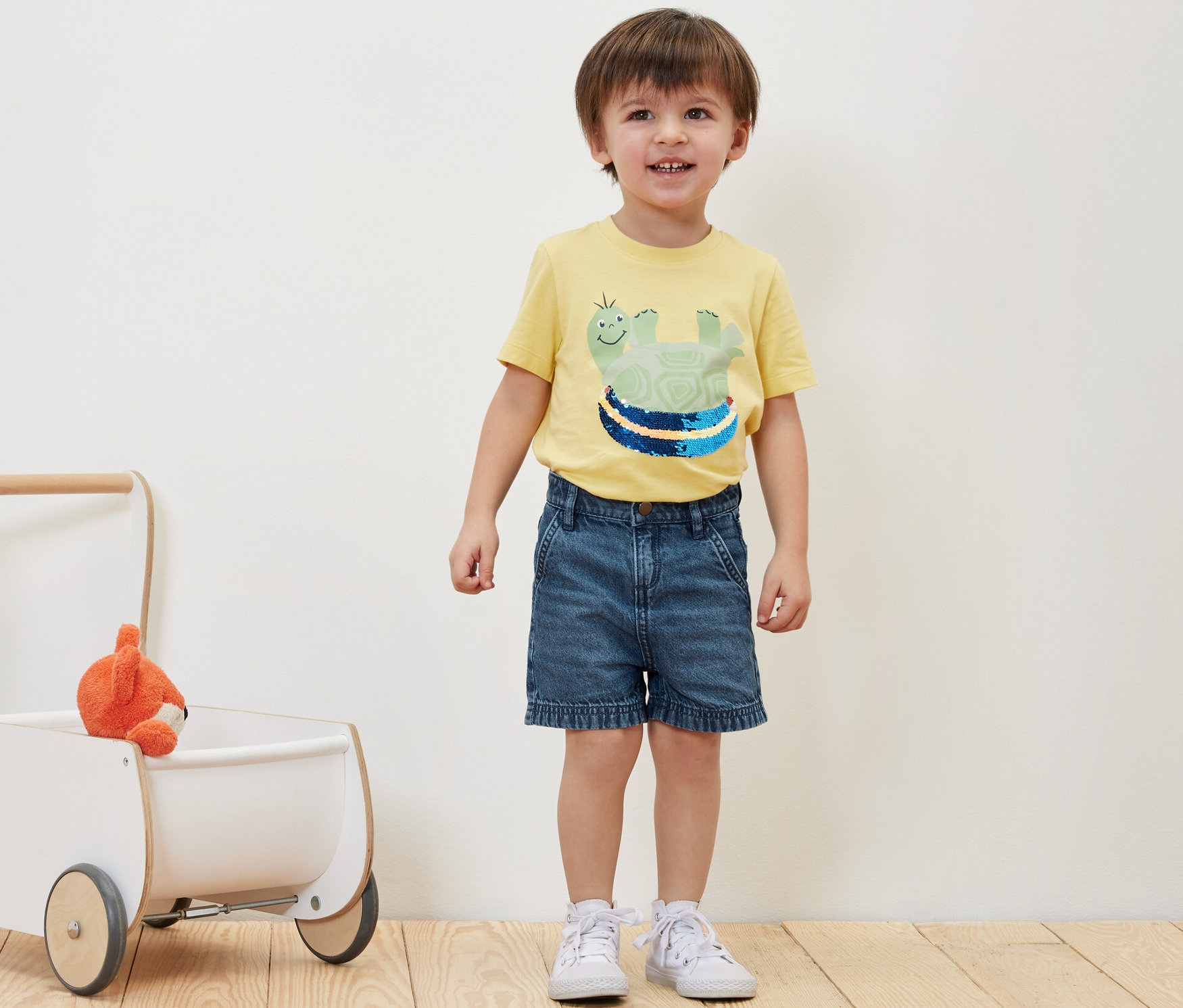 Junge steht neben Kinder-Jeansshorts und Spielzeug im Lauflernwagen.