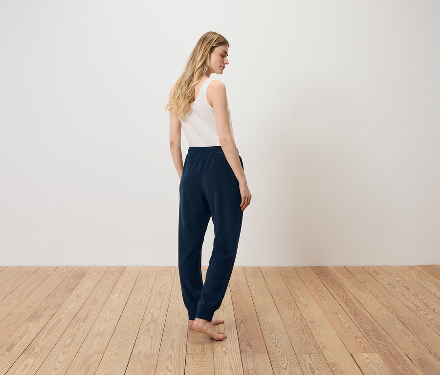 Frau steht mit dem Rücken zur Kamera, trägt ein weißes Tanktop und dunkelblaue Jogginghose.