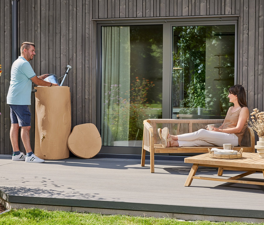 Mann gießt Wasser in einen Holzfass-Regenwassertank, während eine Frau auf einer Holzbank mit Kaffeetasse sitzt.
