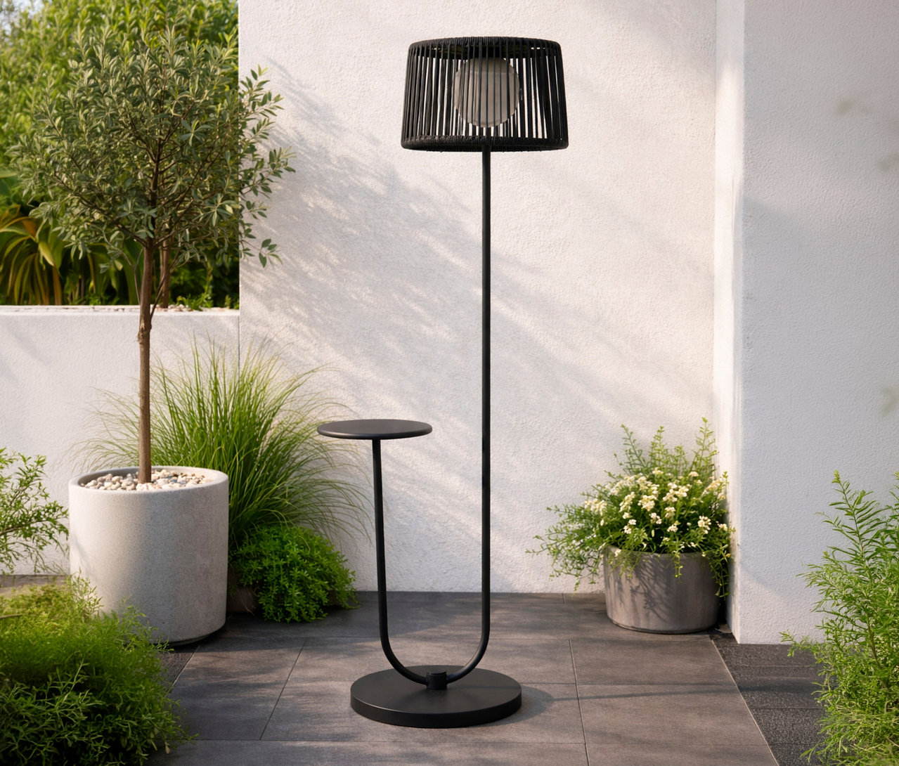 Draußen steht eine anthrazitfarbene Leco Solar Stehleuchte mit integriertem Tisch neben einem Baum und Pflanzen.