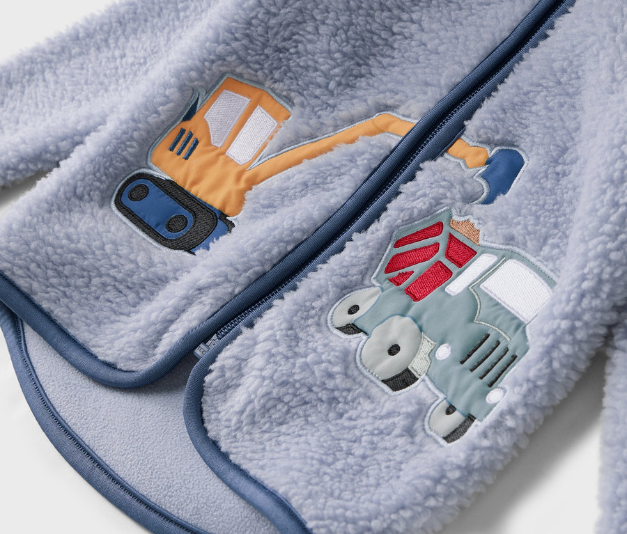 Detailansicht einer hellblauen Fleecejacke mit Reißverschluss und aufgenähten Motiven eines Baggers und eines Trucks.
