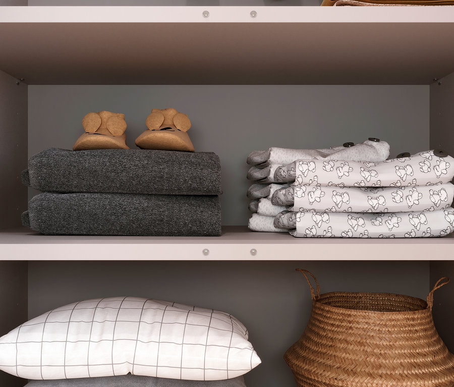 Regal mit zwei grauen gefalteten Handtüchern mit Babyschuhen, einem Stapel weißer Babykleidung mit Bärendruck, einem Kissen mit quadratischem Muster und einem Korb.