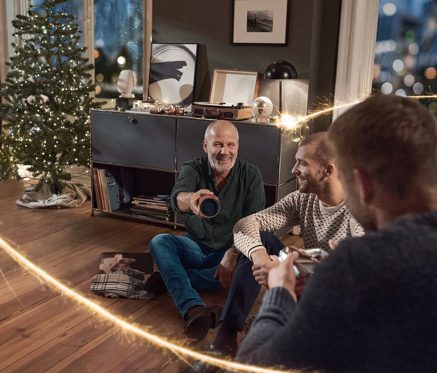 Drei Männer sitzen im Wohnzimmer auf dem Boden, einer hält einen Vintage-Bluetooth®-Lautsprecher mit Kunstlederhülle. Sie tragen Jeans, einen Strickpullover und ein Flanell-Hemd.