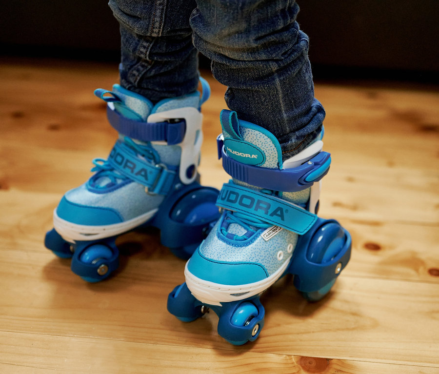 Beine eines Kindes in blauen HUDORA Rollschuhen "My First Quad" auf einem Holzboden.