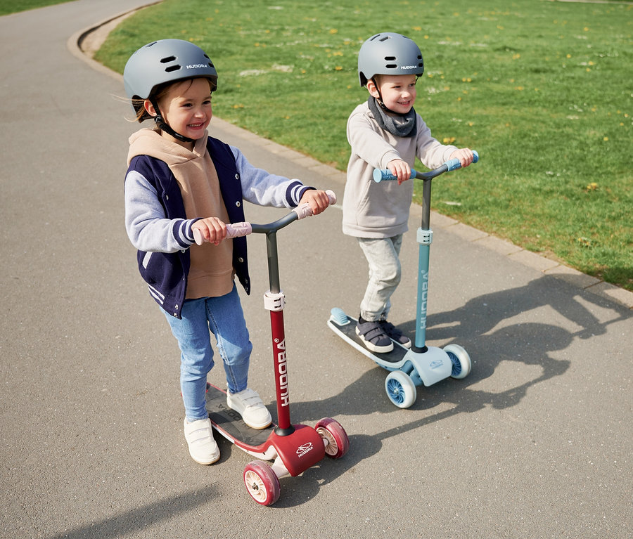 Zwei Kinder mit Helmen fahren auf HUDORA Tri-Scootern auf einem Gehweg, eines bordeauxrot, eines blau.