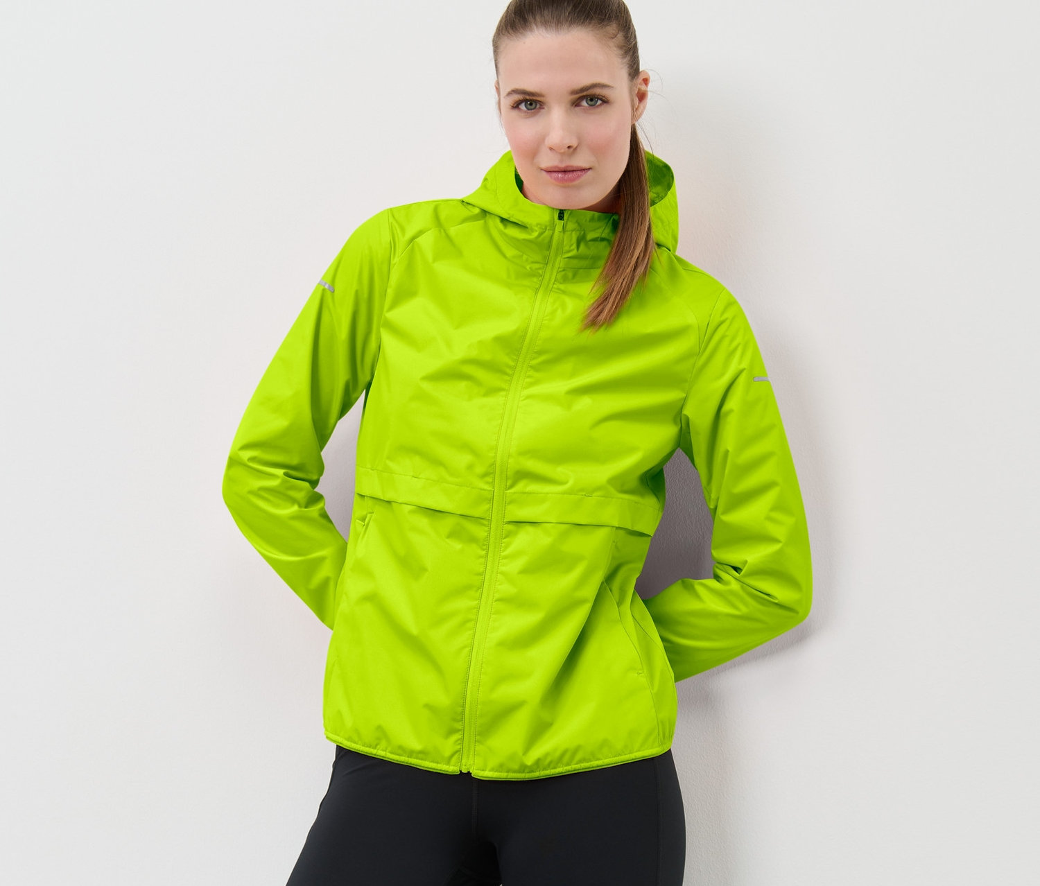 Frau posiert vor weißem Hintergrund in Windprotection-Laufjacke, neonlime und schwarzen Leggings.