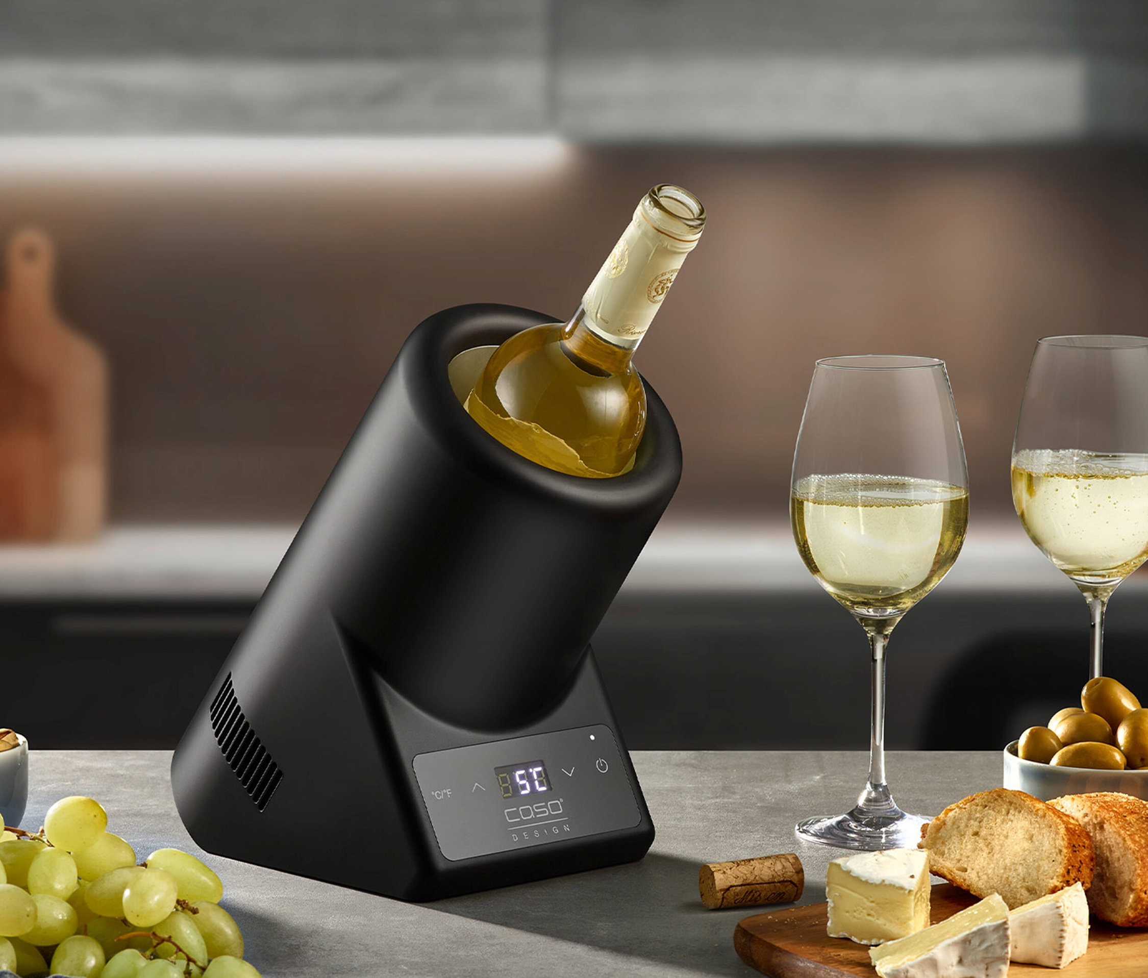 Eine Flasche Weißwein im schwarzen CASO Weinkühler VinoCase neben zwei Gläsern Wein, Käse und Trauben.