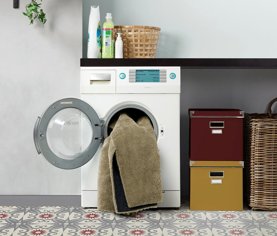 Ein beiger Teppich »Mellow« liegt im Waschraum neben einer Waschmaschine.