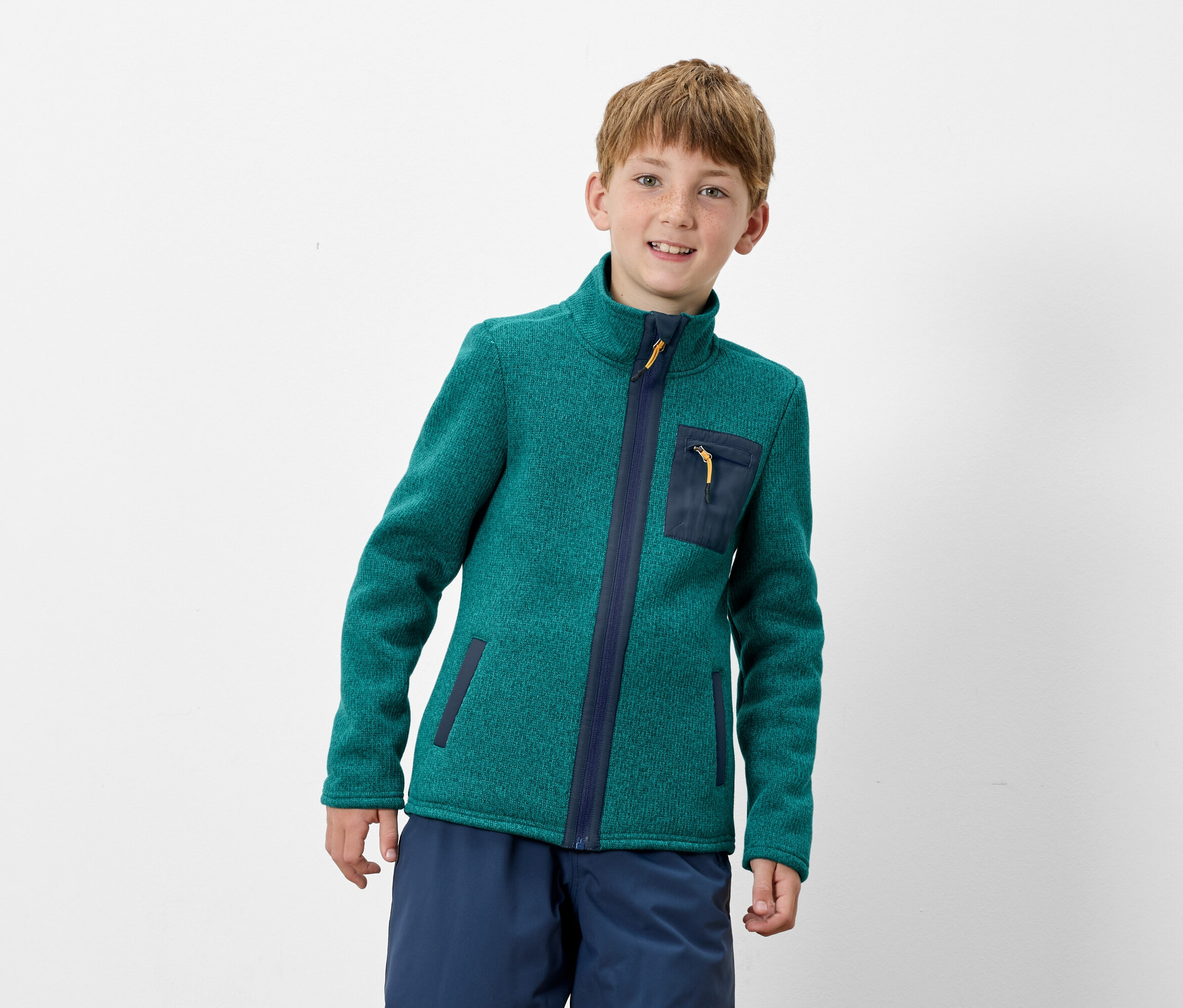Ein Junge trägt eine grüne Fleecejacke mit Reißverschluss und dunkelblauen Details und blaue Hosen.