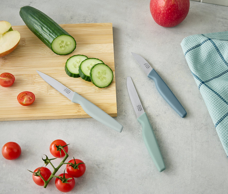 Auf einem Holzbrett liegen eine halbe Apfel, eine Gurke und geschnittene Gurkenscheiben. Daneben liegen drei Messer und Cherrytomaten.